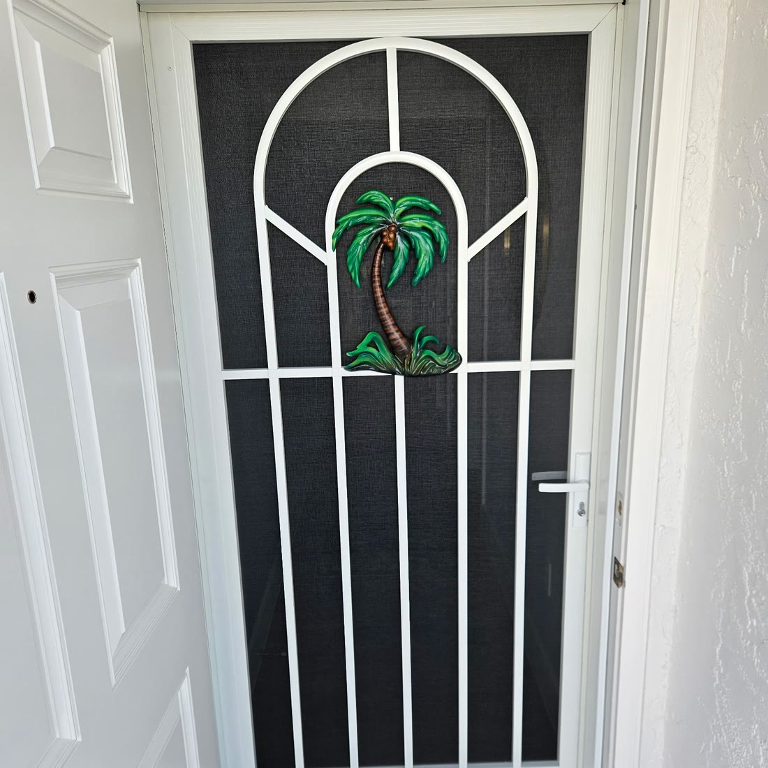 A white aluminum screen door with an arched grid and a palm-tree insert, beside a white front door.