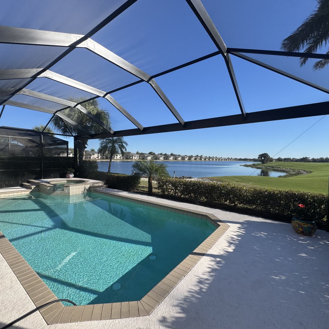 Inside a dark bronze pool cage: a pool and spa, a lake and a golf course beyond the screen.