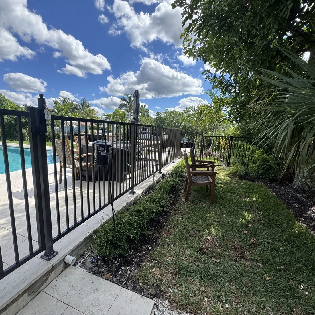 A black aluminum railing along the edge of a pool deck, with patio chairs on the lawn behind it.