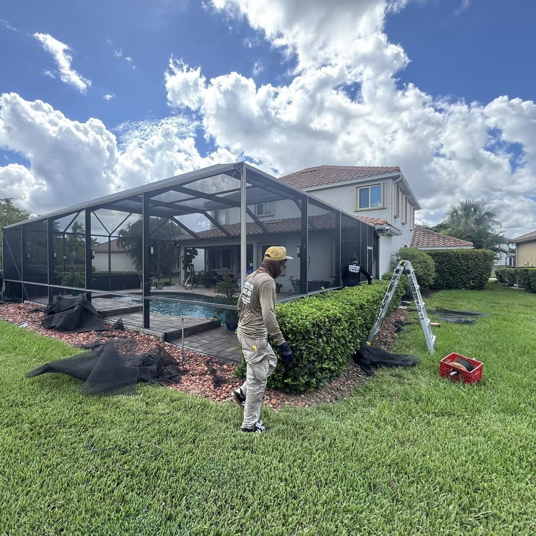One of the crew walking past a hedge beside a pool cage, old screen rolled on the grass and a ladder by the cage.
