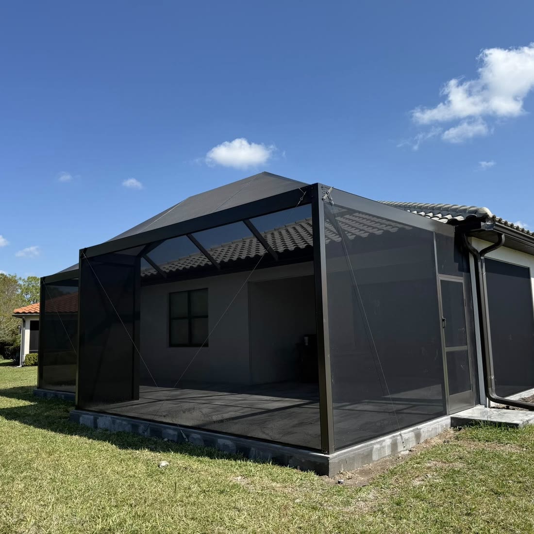 A new bronze screen room on the back of a grey house, on a green lawn under a blue sky.