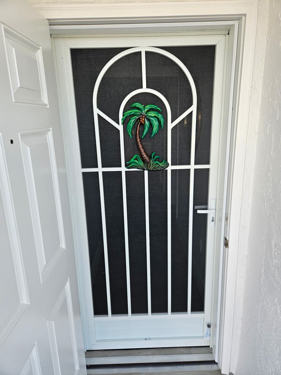 A white aluminum screen door with an arched grid and a palm-tree insert, beside a white front door.