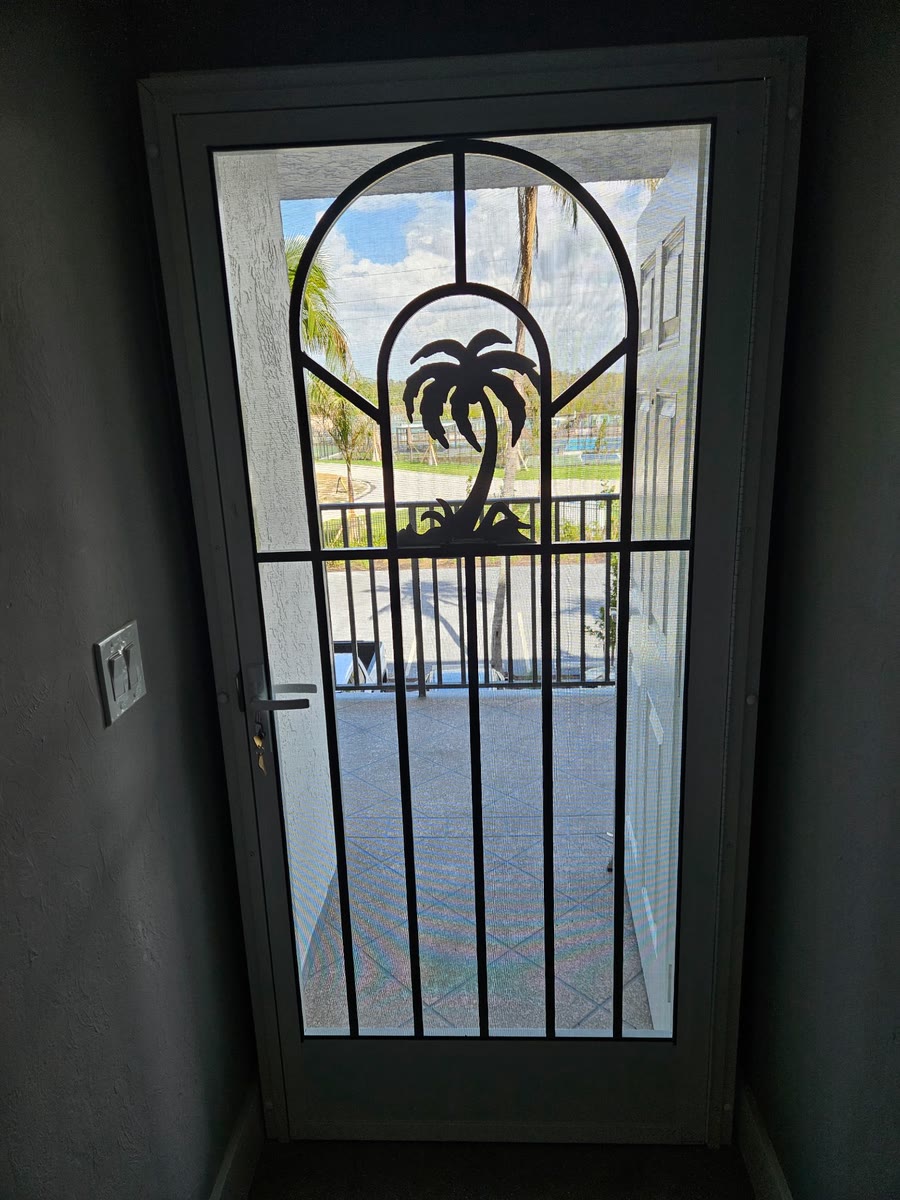 A dark screen door with an arched grid and a palm-tree insert, a lake showing through it.
