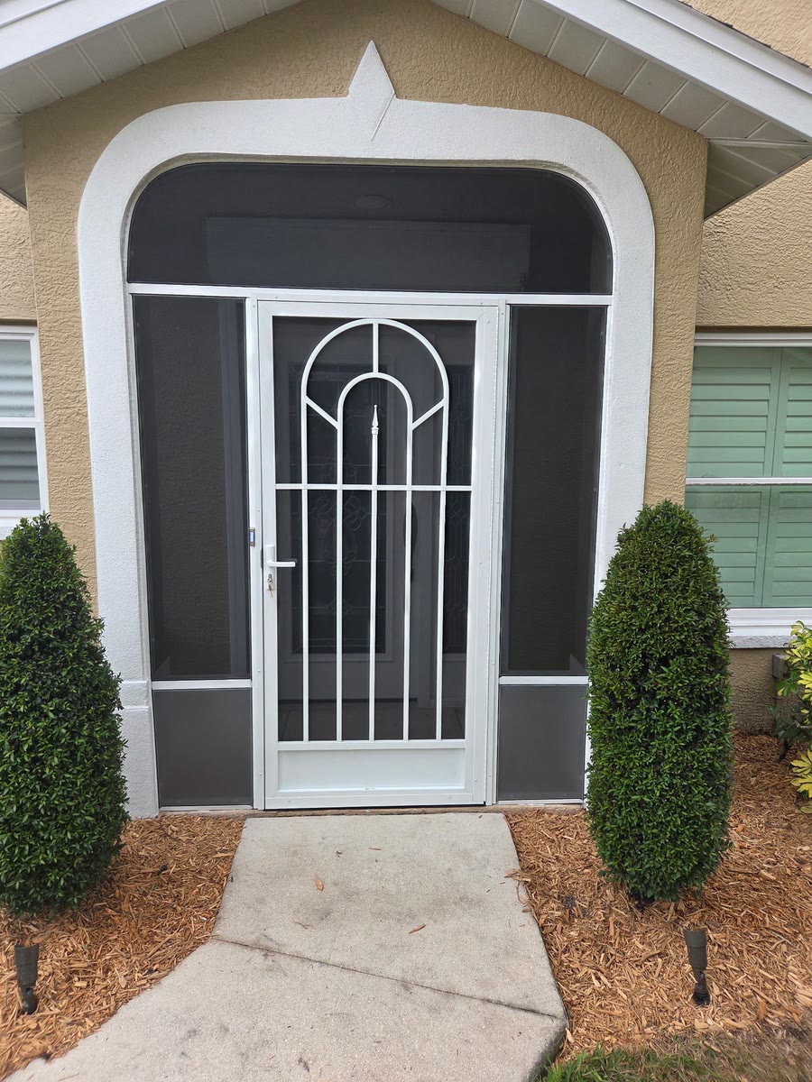 A white screen door with an arched grid in a front entry, two shrubs on either side.