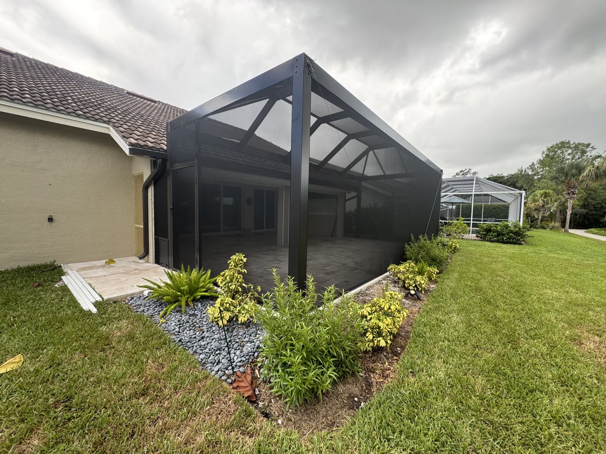 A new bronze pool cage beside a cream house, with fresh landscaping along it.
