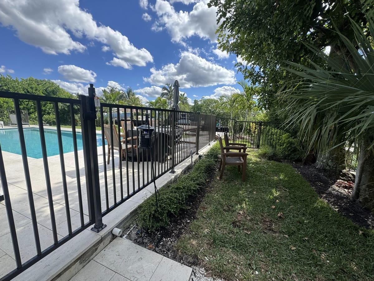 A black aluminum railing along the edge of a pool deck, with patio chairs on the lawn behind it.