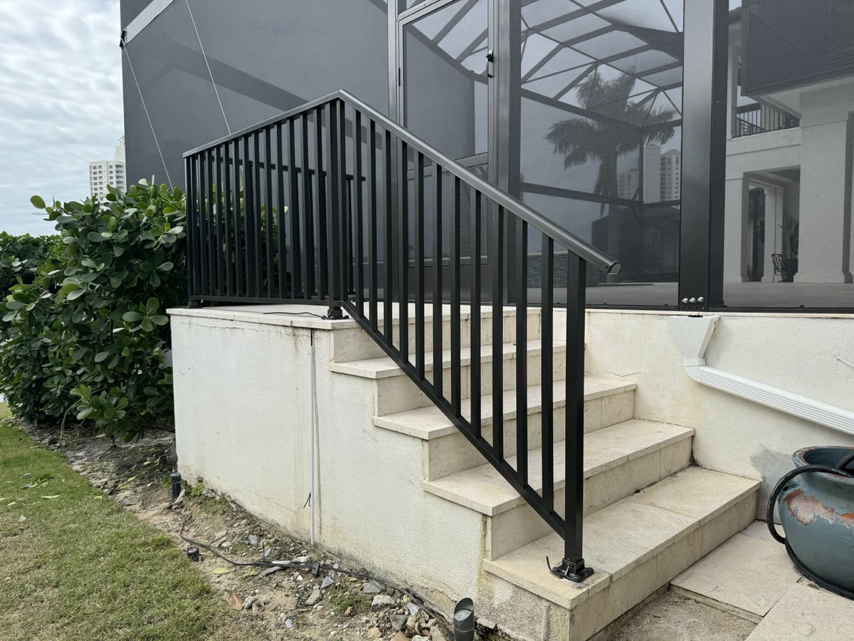 A black aluminum handrail up a set of white concrete steps beside a pool cage.