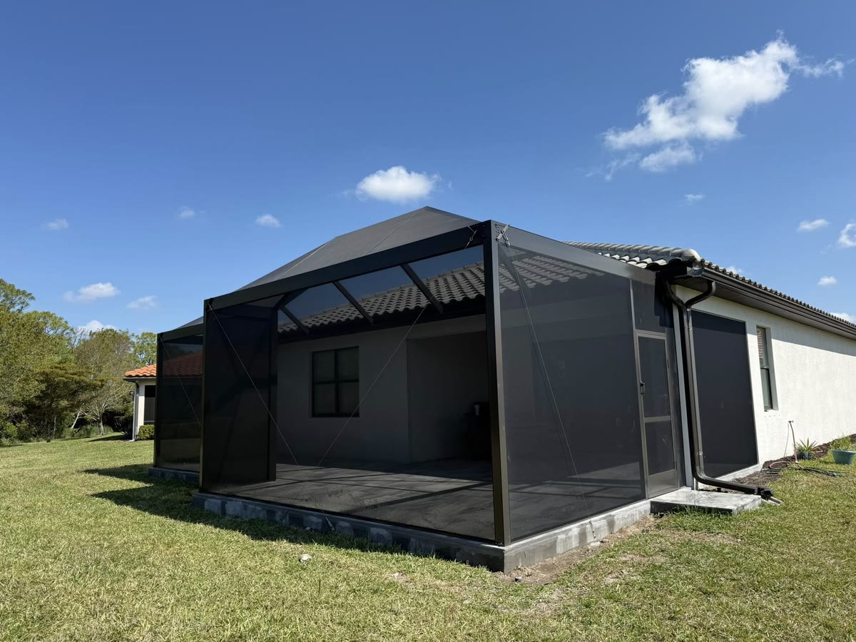 A new bronze screen room on the back of a grey house, on a green lawn under a blue sky.