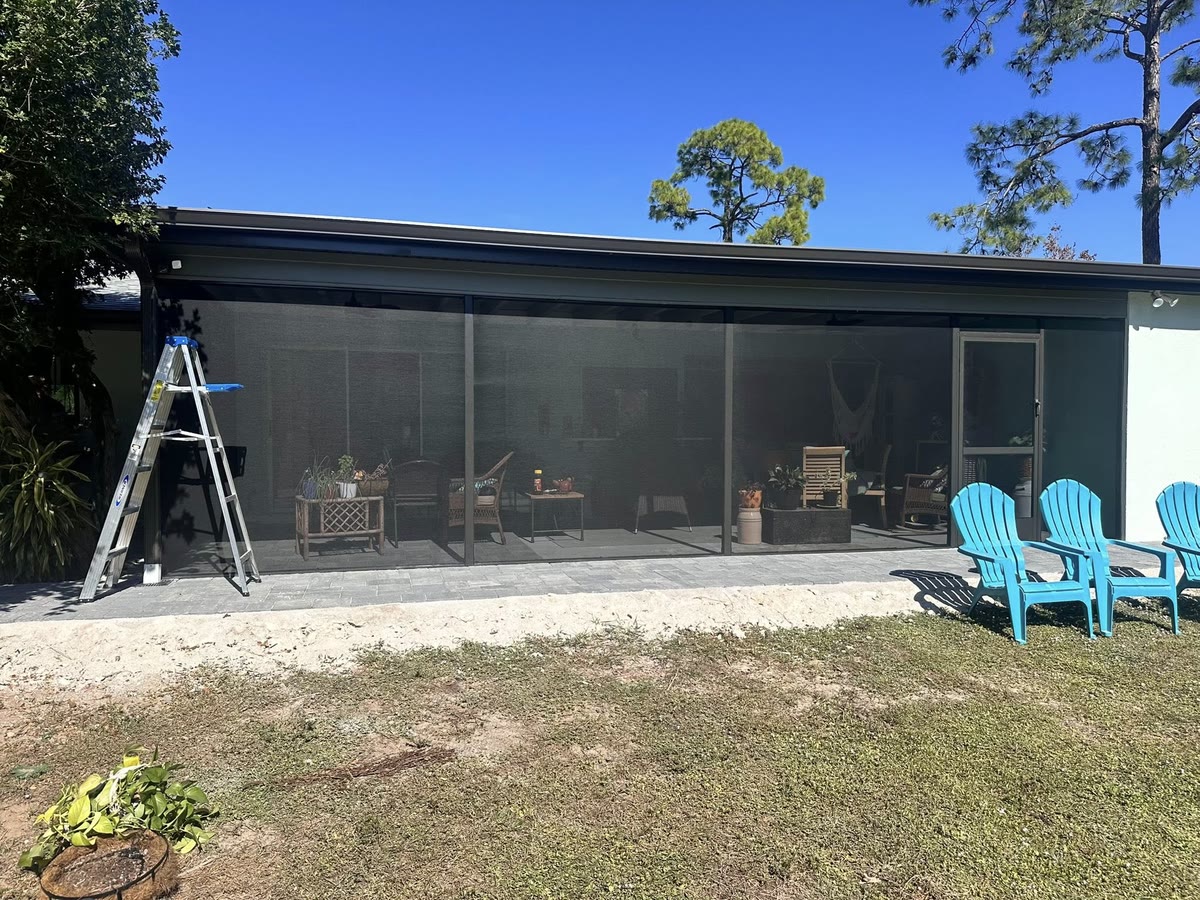 A long screen room across the back of a house, a ladder and blue chairs outside.