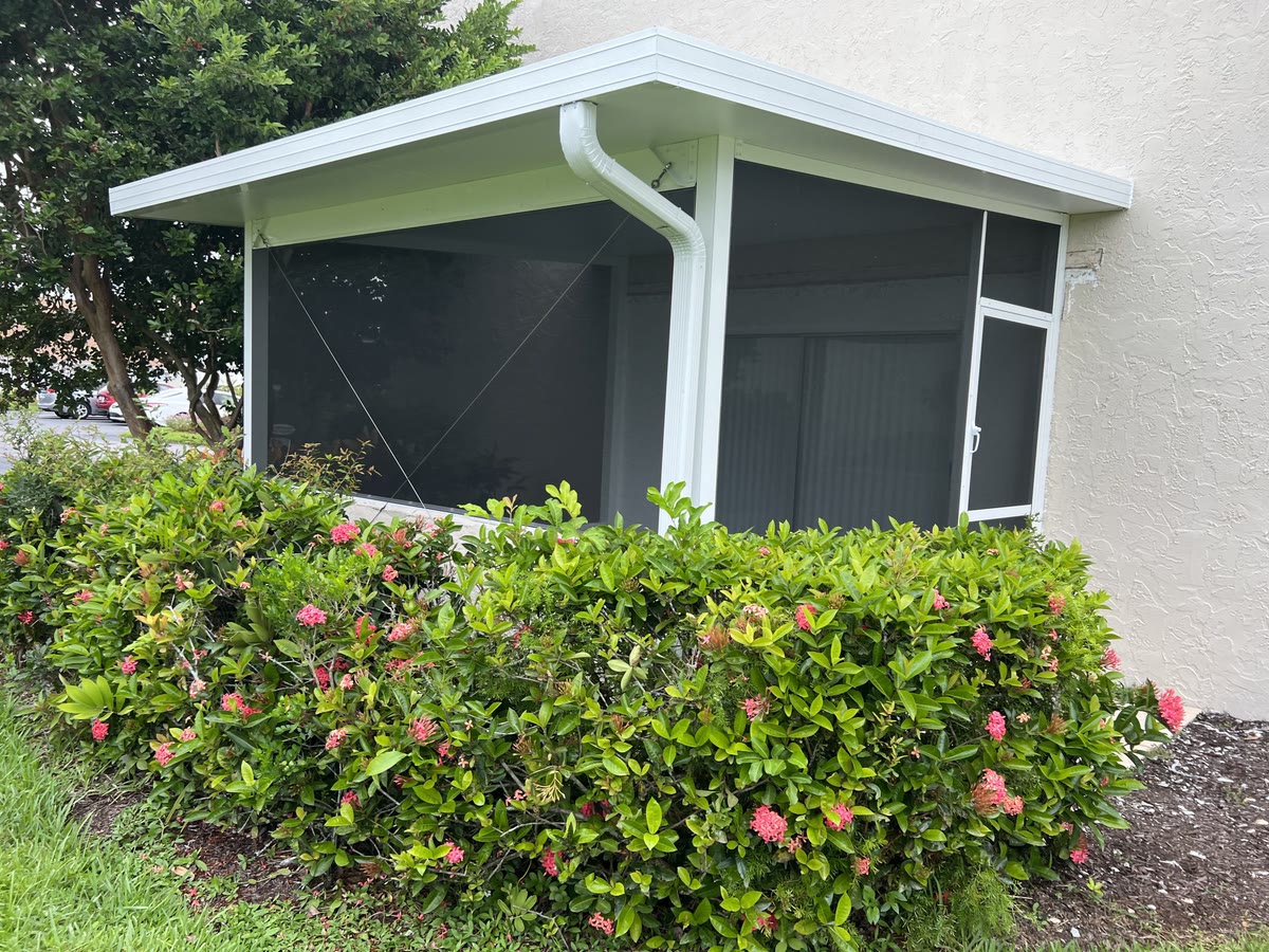 A small white screen room with a flat roof behind flowering shrubs.
