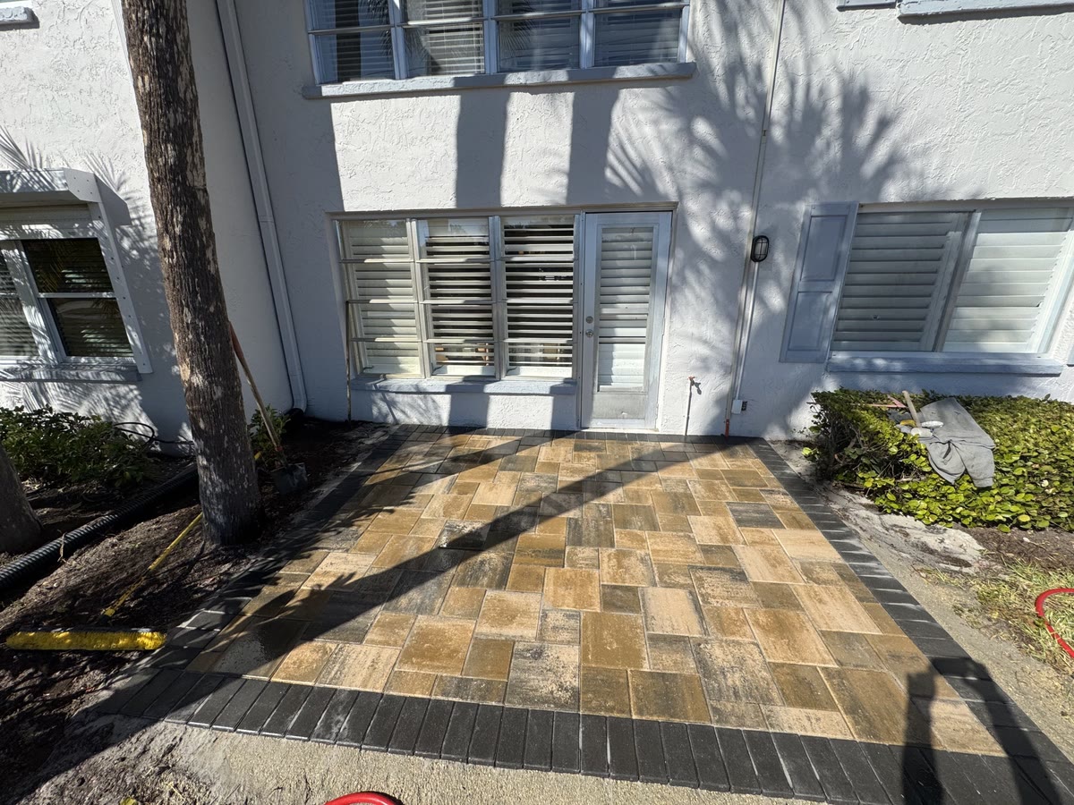 A new patio of tan and grey pavers below a white house with shuttered windows.